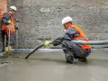 Dwóch budowlańców w kamizelkach odblaskowych wylewa pianobeton. Jeden kieruje wąż, drugi sprawdza poziom.