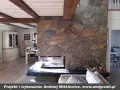 Kamienny kominek z naturalnego łupku, z drewnianymi belkami na suficie, tworzy przytulną atmosferę w salonie.