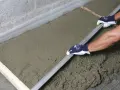 Ręce w rękawiczkach wyrównują świeży styrobeton za pomocą łaty.