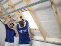 Dwóch fachowców montuje stelaż pod płyty gipsowo-kartonowe, przygotowując ocieplenie dachu. Jeden z nich używa poziomicy, drugi wkrętarki.