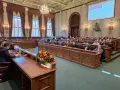 Sala obrad z ludźmi siedzącymi w ławach, przed nimi drewniane biurka, na jednym z nich bukiet kwiatów.