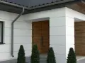 Nowoczesny dom z białym boniowaniem, drewnianymi drzwiami i ozdobnymi tujami.