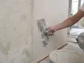 Ręka z kielnią wygładza ścianę podczas gipsowania. Widać starą, łuszczącą się farbę i wiadro z gipsem.