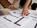 Dłoń wskazująca na plan mieszkania, obok laptop i próbki materiałów.