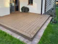 Nowoczesny taras z deski kompozytowej, otoczony zieloną trawą i kamienną ścieżką, tworzy idealne miejsce do relaksu.