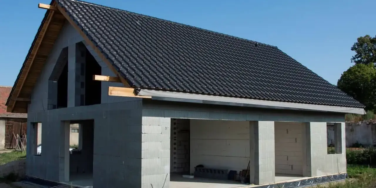 Dom w stanie surowym otwartym, z dachem pokrytym czarną dachówką. Ściany z bloczków betonowych, widoczne otwory okienne i drzwiowe.