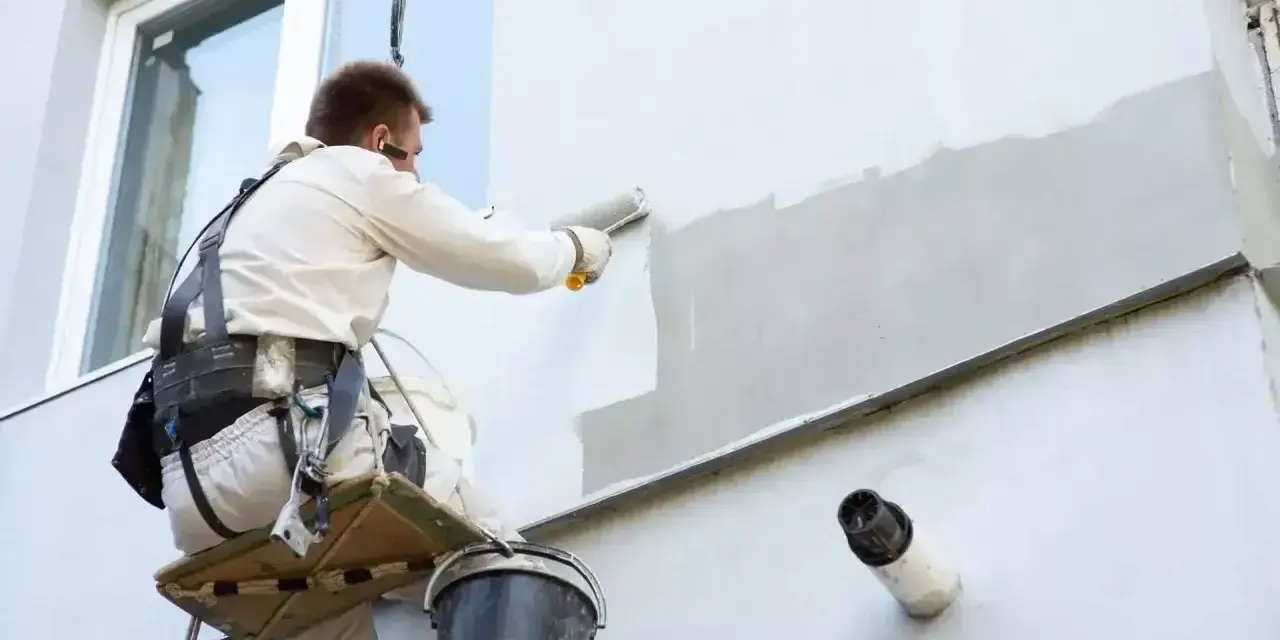 Malowanie elewacji cena: pracownik na wysokości maluje ścianę budynku wałkiem.