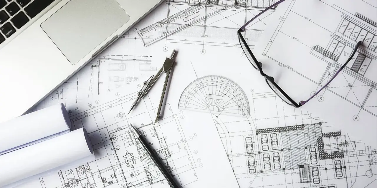 Na stole leżą plany architektoniczne, laptop, okulary i przybory kreślarskie. To miejsce pracy nad projektem i jego kosztorysem.