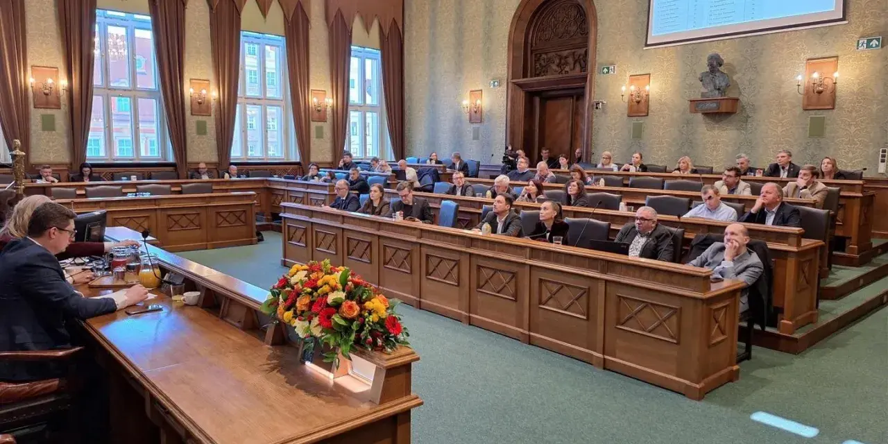 Sala obrad z ludźmi siedzącymi w ławach, przed nimi drewniane biurka, na jednym z nich bukiet kwiatów.