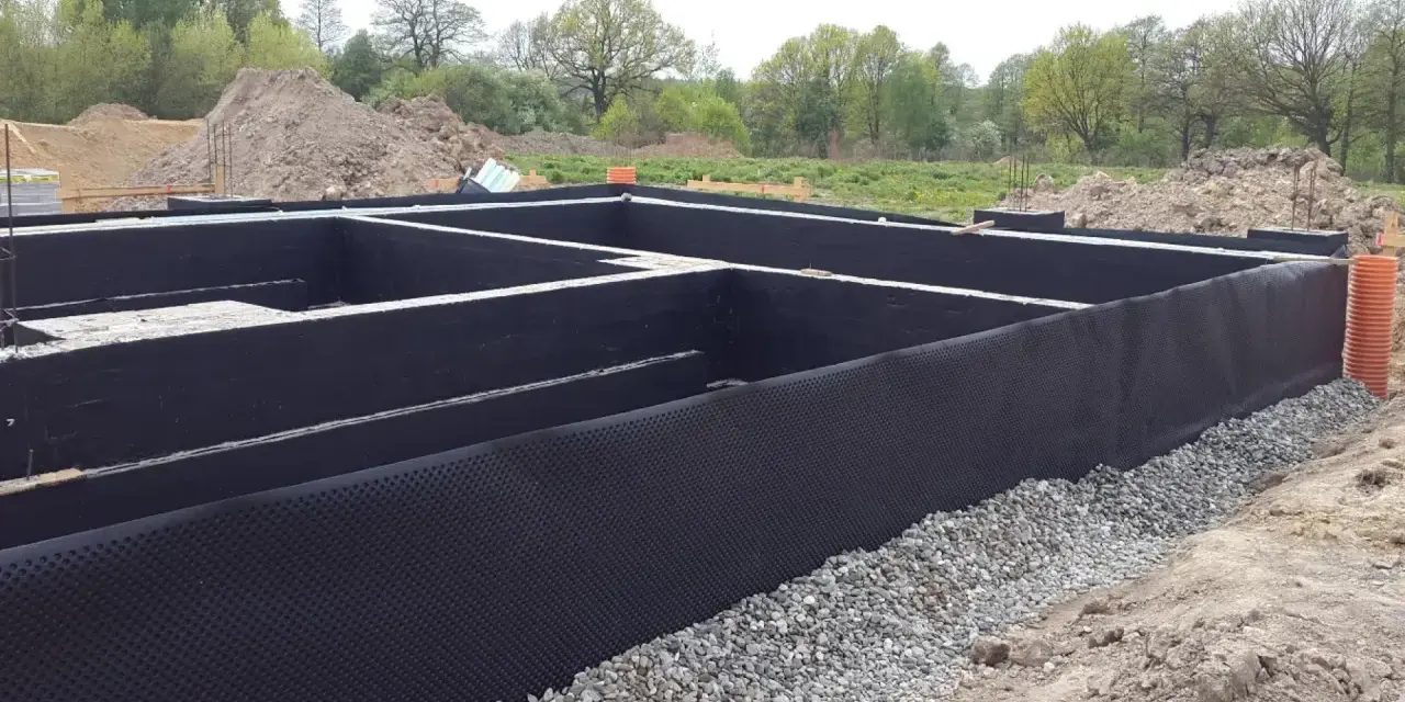 Fundament domu w trakcie budowy, z czarną membraną i otoczakami. Trwa ocieplenie fundamentów, przygotowanie do dalszych prac.