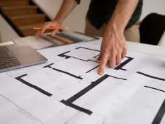 Dłoń wskazująca na plan mieszkania, obok laptop i próbki materiałów.