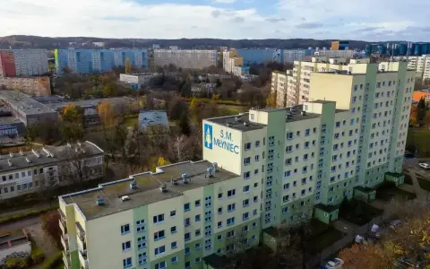 Duży blok mieszkalny S.M. Młyniec z charakterystycznym logo, otoczony innymi budynkami i drzewami jesienią.