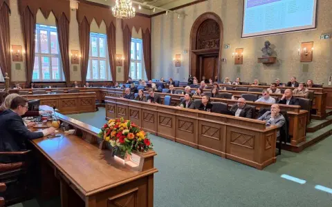 Sala obrad z ludźmi siedzącymi w ławach, przed nimi drewniane biurka, na jednym z nich bukiet kwiatów.