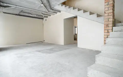 Wnętrze w stanie deweloperskim: betonowe schody, nieotynkowane ściany, widoczne instalacje i przewody.