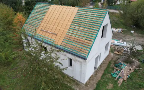 Rozbudowa domu w toku. Dom z białego pustaka, na dachu widoczna więźba i łaty. Wokół materiały budowlane.