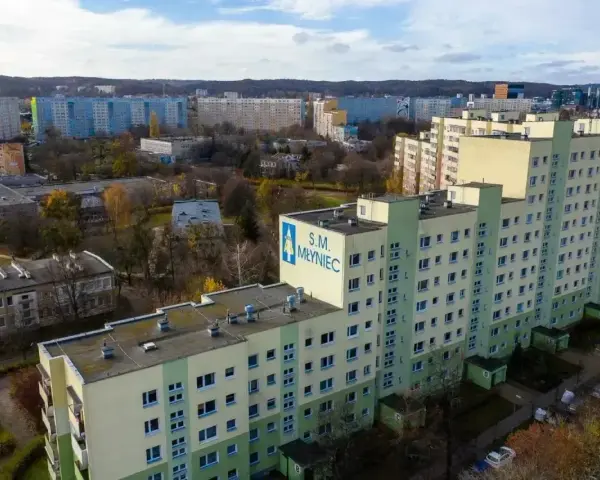 Duży blok mieszkalny S.M. Młyniec z charakterystycznym logo, otoczony innymi budynkami i drzewami jesienią.