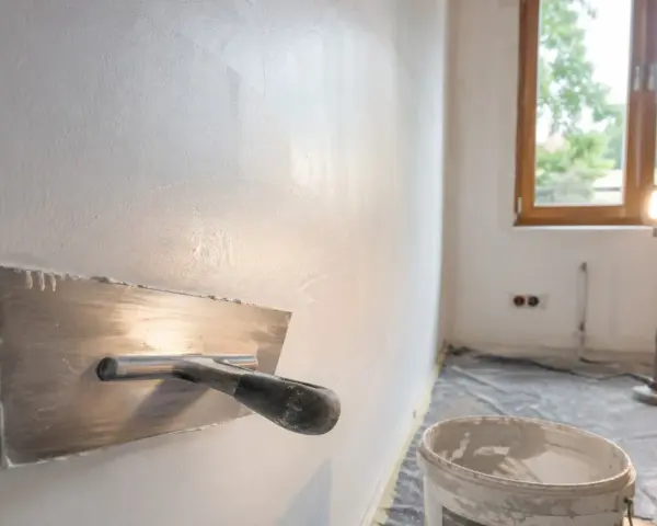 Nakładanie gładzi na ścianę za pomocą kielni. W tle okno i lampa budowlana.