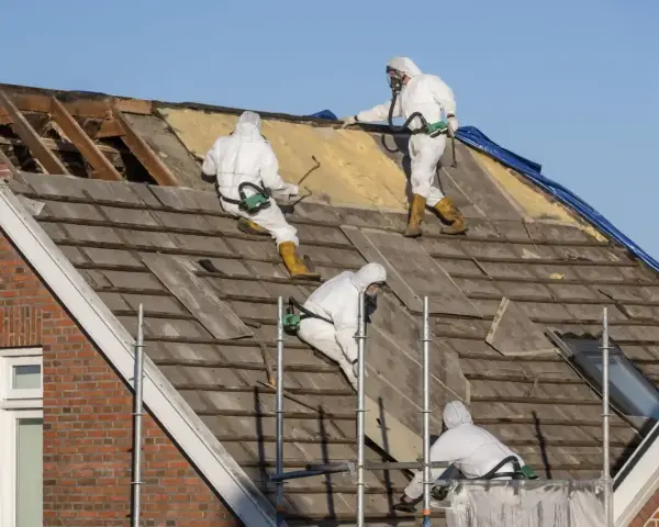Pracownicy w kombinezonach ochronnych usuwają stare pokrycie dachowe, potencjalnie zawierające azbest.