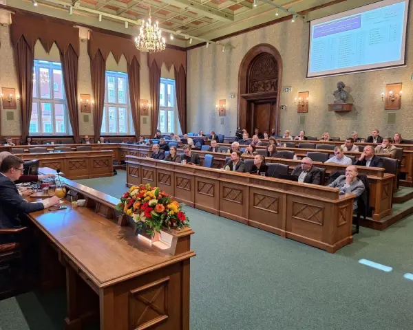 Sala obrad z ludźmi siedzącymi w ławach, przed nimi drewniane biurka, na jednym z nich bukiet kwiatów.