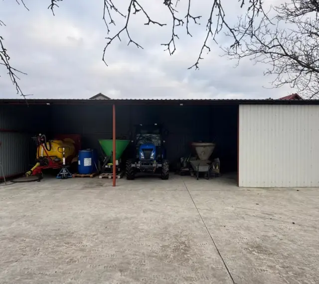 Nowa wiata bez pozwolenia chroni niebieski traktor, żółty opryskiwacz i inne maszyny rolnicze.