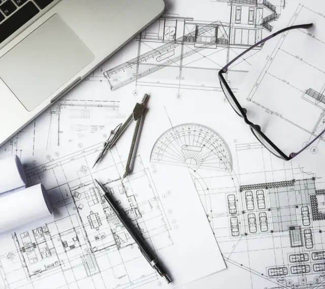 Na stole leżą plany architektoniczne, laptop, okulary i przybory kreślarskie. To miejsce pracy nad projektem i jego kosztorysem.