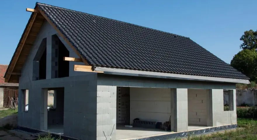 Dom w stanie surowym otwartym, z dachem pokrytym czarną dachówką. Ściany z bloczków betonowych, widoczne otwory okienne i drzwiowe.