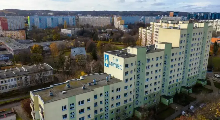 Duży blok mieszkalny S.M. Młyniec z charakterystycznym logo, otoczony innymi budynkami i drzewami jesienią.