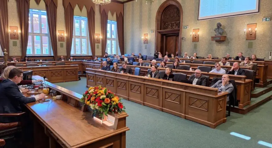 Sala obrad z ludźmi siedzącymi w ławach, przed nimi drewniane biurka, na jednym z nich bukiet kwiatów.