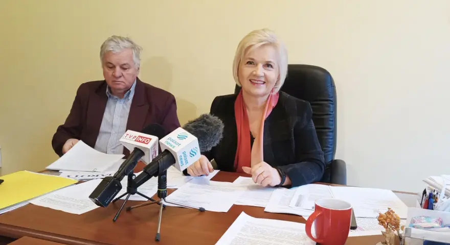 Dwoje ludzi przy biurku z mikrofonami i dokumentami.