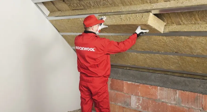 Pracownik w czerwonym kombinezonie Rockwool montuje izolację na ścianie szczytowej poddasza.