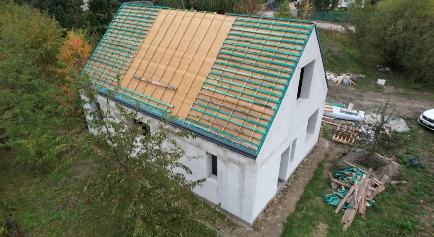 Rozbudowa domu w toku. Dom z białego pustaka, na dachu widoczna więźba i łaty. Wokół materiały budowlane.