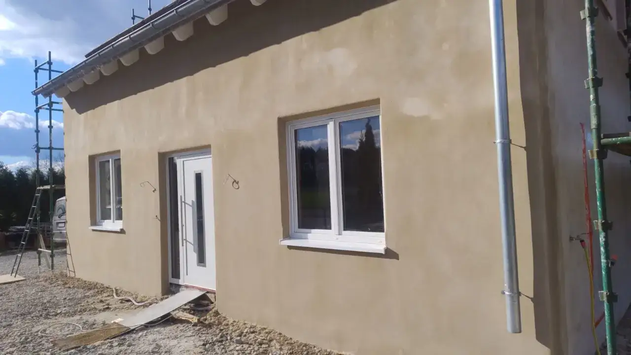 Ściana domu ocieplona tynkiem wapno gaszone, z białymi oknami i drzwiami. W tle rusztowania i niebo.