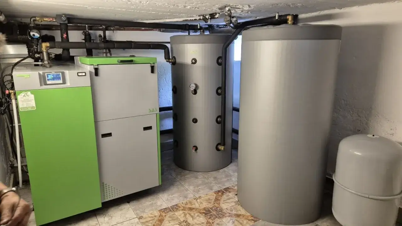 Zielony kocioł i dwa szare bojlery tworzą system grzewczy. Jeden z bojlerów to bufor ciepła, który gromadzi nadwyżki energii.