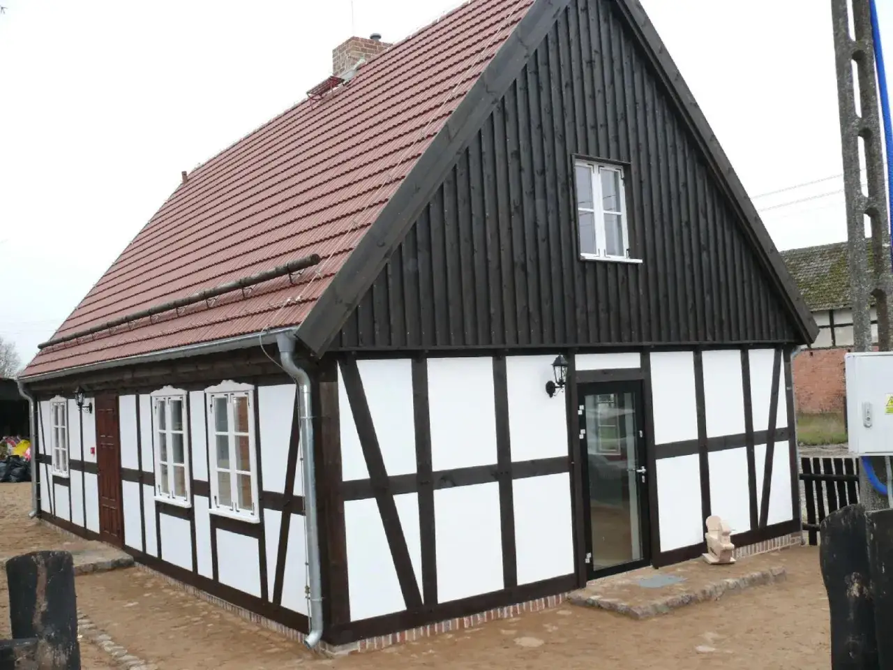 Dom w stylu mur pruski z ciemną drewnianą szalówką i białymi wypełnieniami. Dach kryty czerwoną dachówką.