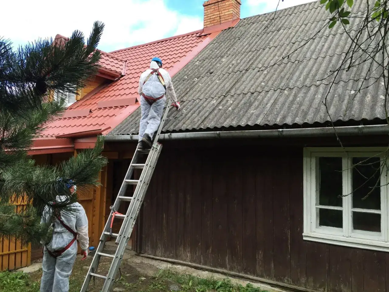 Pracownicy w kombinezonach ochronnych usuwają eternit z dachu. Stary dach pokryty eternitem wymaga specjalistycznego demontażu.