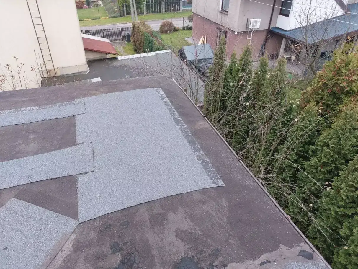 Widok z góry na dach pokryty papą. Widać fragmenty budynków i drzew.